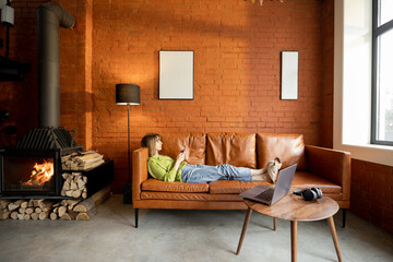 Woman using phone while relaxing on a leather couch at home. Modern Living room with fireplace in loft style. Home coziness and leisure time with gadgets