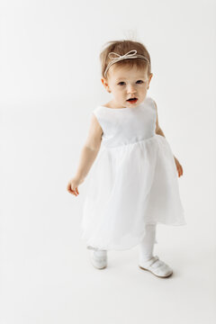 Little blonde positive princess girl in white casual dress and shoes standing walking with over white wall background. Stylish comfortable everyday fashion for children concept