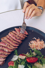 Woman's hand holding fork and eating beef steak with salad, in cafe.