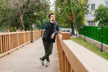 Handsome man wearing denim clothes leaning to wooden fence and looking away while taking on phone with friends or girlfriend.