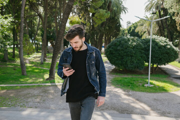 Young man wearing jeans surfing on phone or reading message while walking on city park. Man typing a message to girlfriend.