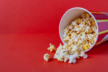 popcorn spills out of a striped paper bucket on a red background, copy space