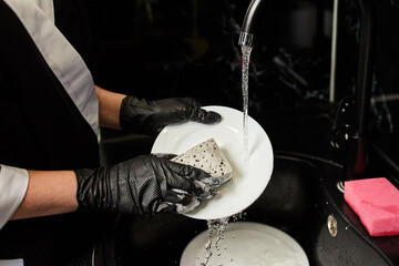 Professional Cleaning service company employee in rubber gloves washes dishes in the sink on the kitchen close up, disinfects and sterilizes