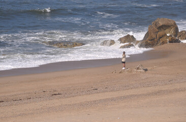 Pessoa do sexo feminino parada em cima de uma rocha numa praia a observar o oceano - obrazy, fototapety, plakaty