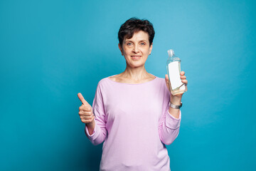 Happy friendly mature woman holding cleaning bottle and showing thumb up on blue background
