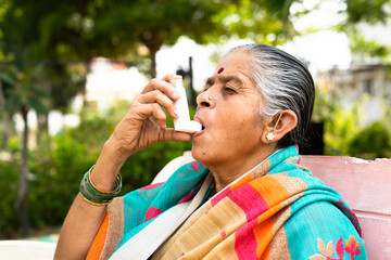 elderly woman using asthma inhaler while sitting at park due to allergy - concept showing effects of pollution, illnes and disease - obrazy, fototapety, plakaty
