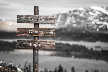 let the games begin text quote written on wooden signpost outdoors in nature with lake and mountain scenery in the background. Moody feeling.