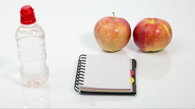 Two red ripe apples, a bottle of water, a pen and a notebook with the handwritten word diet on a white table. The concept of weight loss, organic products, healthy diet planning. Stop motion animation