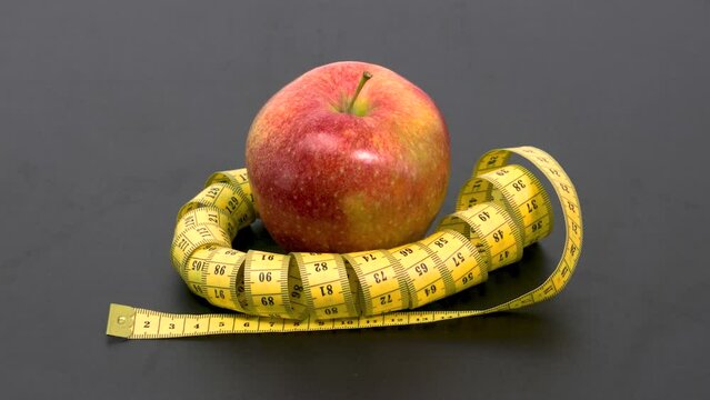Red ripe apple wrapped in swirls of yellow measuring tape on a black background. The concept of weight loss, healthy eating, diet. Stop motion animation.
