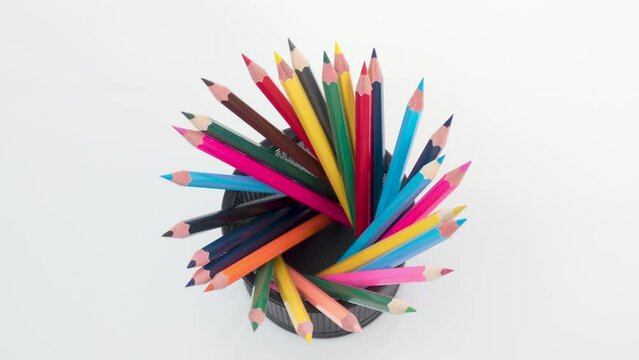 A black metal holder on a white table is filled with colorful wooden pencils. Variety, rainbow colors, stop motion animation.