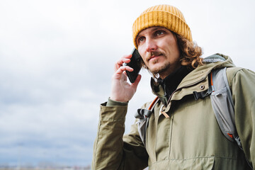 A guy in a yellow hat and jacket is talking on the phone on the street, a hipster holds a smartphone in his hand against the sky, a tourist on a trip communicates with loved ones.