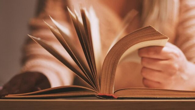 Young unrecognizable woman slowly turning the pages of an old thick book on the table, studying in the library, reading, searching for information, leisure at home.