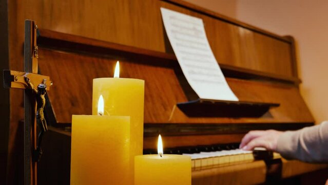 Female musician pianist finishes playing a beautiful religious christian melody on a brown upright piano with musical notes sheet in cozy room or curch with three burning candles and a crucifix and cl