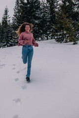 A young, slender, long-haired girl runs through the snow in a winter forest in the mountains.