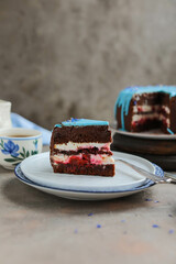 A piece of beautiful chocolate cake with blue icing and cornflower flowers. Biscuit cake with curd cream and cherries on a table with tea utensils.