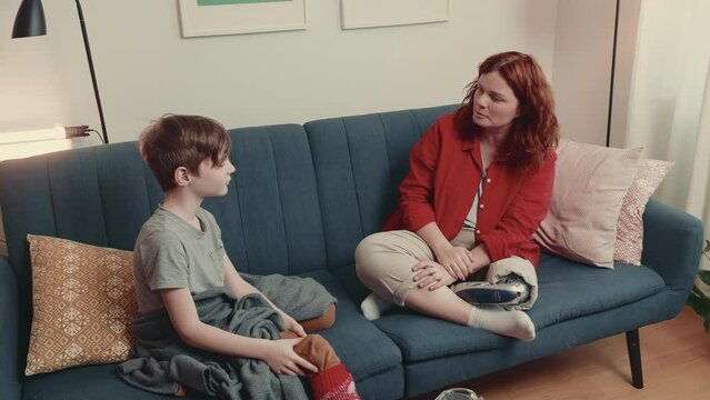 Beautiful happy disabled tutor woman with bionic prosthetic leg talks to teenage boy child sitting on couch at home.