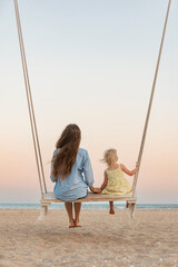 Young mother and little blond girl sit on swing on the beach during sunset. Mom and daughter on...