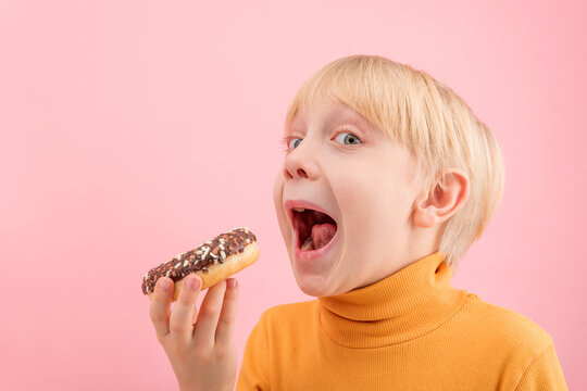 Towheaded boy wants to bite donut with chocolate icing. Close up portrait of child with doughnut on pink background.