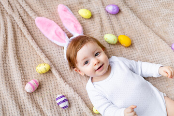 cute little baby with bunny ears and painted eggs lies on a beige background and smiles, easter concept