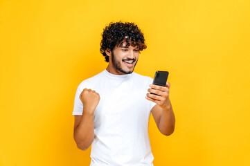 Fototapeta Happy joyful excited Indian or Arabian guy holds smartphone, get unexpected news, winning lottery, stands on isolated orange background, cheerful facial expression, toothy smile, gesturing with fist obraz