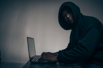 A hacker wears a long-sleeved shirt with a hood covering his head. Sitting in a dark room using a laptop The concept of identity theft on the Internet camp network. with copy space.