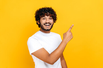 Cheerful indian or arabian guy in white basic t-shirt, looks and points finger to the side at mock-up space for your presentation, stands on isolated orange color background, looks at camera, smiling