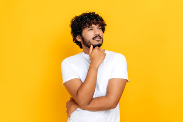 Portrait of a handsome optimistic indian or arabian guy in basic t-shirt, looking pensively to the side at empty space, thinking new ideas, stands on isolated orange background, holds hand near chin