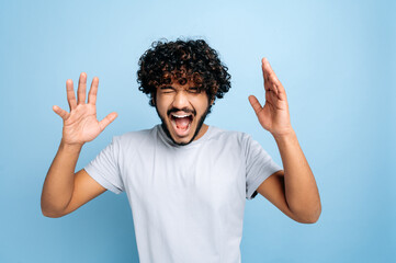 Annoyed curly-haired indian or arabian guy in a t-shirt, shouting loudly with his eyes closed,...