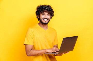 Attractive contented indian or arabian guy in t-shirt, holding an open laptop in his hand, looks friendly at the camera while standing against isolated orange background, smiling happily