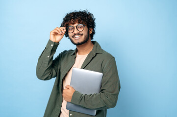 Smart handsome positive indian or arabian millennial guy, with glasses, student or freelancer, holding a laptop in hand, standing on isolated blue background, looking at the camera, smiling friendly - obrazy, fototapety, plakaty