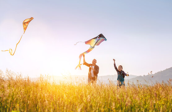 Summer outdoor photo of smiling father with daughter as they releasing colorful kites on the high grass meadow. Warm family moments or outdoor time spending concept image.