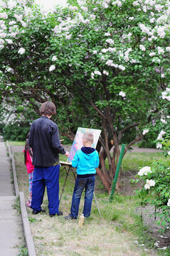 painter with children standing in front of lilac bushes and paints a picture on an easel. Teacher and students in the open air