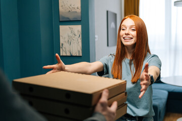 Cheerful excited young woman receiving paper boxes with pizza from unrecognizable courier male on doorway at home. Rear view of delivery man delivering boxes with food to female client at apartment.