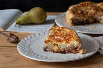 A slice of pears and chocolate cake. Spoon with chocolate cream at the side. Background: a pear on a cookbook.