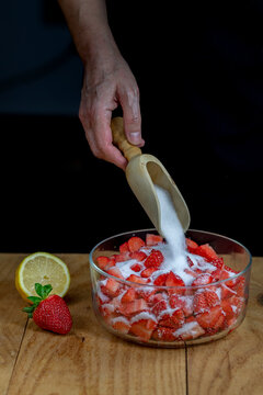 A female hand pouring white sugar on a glass bowl with strawberries with a chadwick wooden spoon.