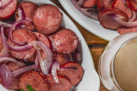 Linguiça frita acebolada com cebola roxa na prato com limão e acompanhada de pão fatiado e cerveja em mesa de madeira no bar ou boteco