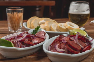 Linguiça frita acebolada com cebola roxa na prato com limão e acompanhada de pão fatiado e cerveja em mesa de madeira no bar ou boteco