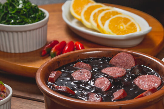 Feijoada com feij&atilde;o preto em tigela de barro com lingui&ccedil;a calabresa acompanhada de arroz branco vinagrete farofa farinha pimenta repolho laranja sobre mesa de bar em madeira