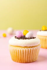Easter vanilla cupcake with cream cheese, colorful candy eggs, chocolate sprinkles on a pink and yellow background. Close-up, copy space