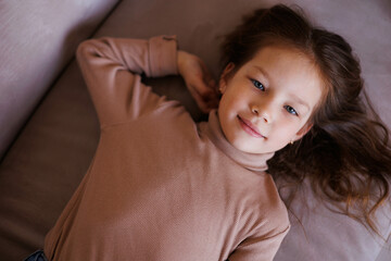 Portrait of 6-year-old little girl laying on her back in casual outfit, on sofa at home. Female child pretty face with beautiful deep eyes. Cute baby girl smiling. Relax and peace. Turtleneck clothes