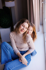 Lovely family mother and daughter embracing and kissing each other indoors. Happy sweet woman with little 6-year-old girl in casual clothers: turtlenecks and jeans sitting on sofa, hugging and smiling