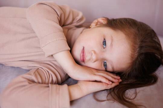Portrait of 6-year-old little girl laying on her back in casual outfit, on sofa at home. Female child pretty face with beautiful deep eyes. Cute baby girl smiling. Relax and peace. Turtleneck clothes