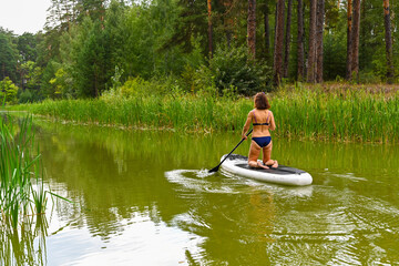 A woman drives on the Sup Board through a narrow canal surrounded by dense grass. Active weekend vacations wild nature outdoor. The woman is standing on the lap in a bathing suit.