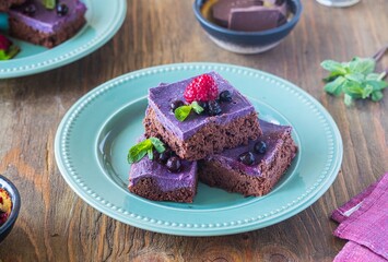 Dessert, sliced chocolate brownies with a layer of ricotta or cream cheese and blueberry puree on a green plate on a brown wooden background. American cuisine.