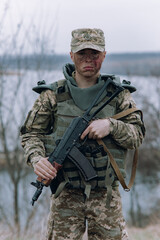 Ukrainian soldier stands with kalashnikov assault rifle in his hands against background of sky and...