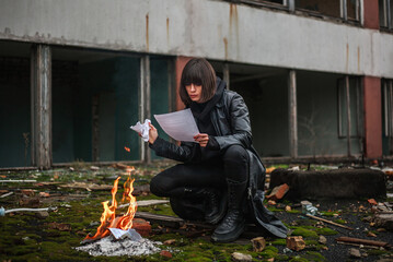 Woman in black leather coat on the background of destroyed and abandoned residential building burns paper documents