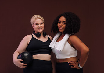 Two cheerful women in sportswear posing on brown background. Plus size females standing together with fitness equipment.