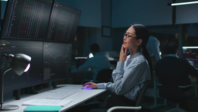 Asian woman trader look at multi-screen workstation with graphs and data working late in dark office