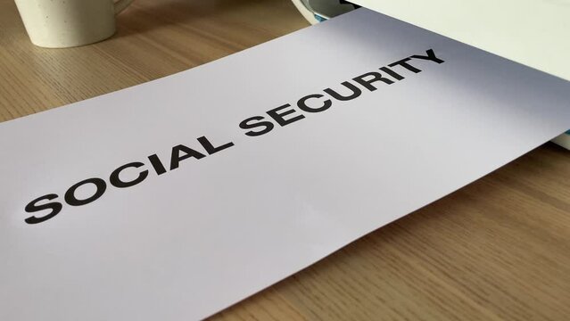 Printing &ldquo;SOCIAL SECURITY&rdquo; text written message word on paper sheet document. Close up A4 page being printed from multifunction scanner printer on wooden desk in office. Hand take paper from printer