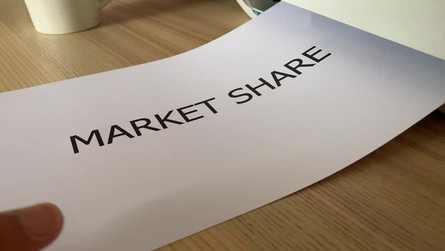 Printing &ldquo;MARKET SHARE&rdquo; text written message word on paper sheet document. Close up A4 page being printed from multifunction scanner printer on wooden desk in office. Hand take paper from printer